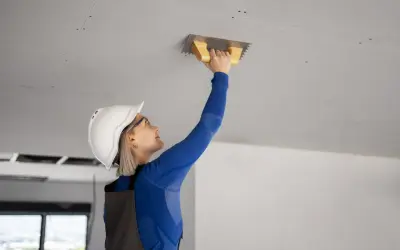 Kobieta w kasku i kombinezonie tynkuje sufit podwieszany kielnią.