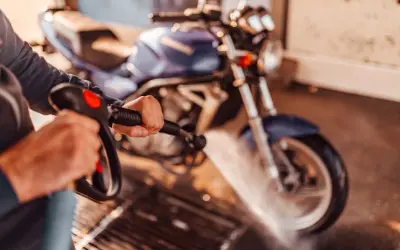 Persona limpiando una moto azul con una hidrolavadora. El agua salpica sobre la rueda delantera.
