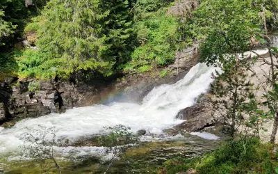 Vattenfall i Idre, med en regnbåge som skimrar i dimman från det forsande vattnet. Omges av grönskande träd och klippor.