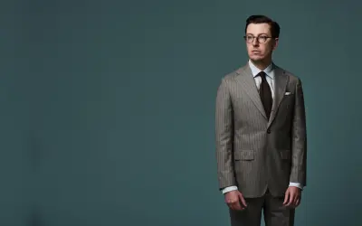 A man in a pinstripe suit, with a brown tie and glasses, stands against a teal background. The jacket length appears to be ideal, hitting just below the seat.