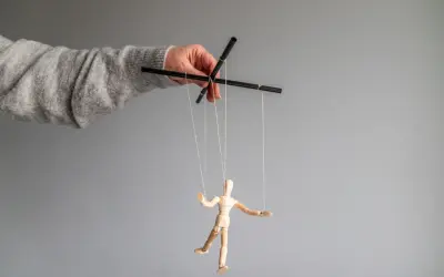 Dłoń w szarym swetrze steruje drewnianą marionetką. To obraz manipulatora, który pokazuje swoje cechy.