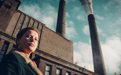 Kobieta na tle dymiących kominów huty Szopienice. Ołowica unosi się w powietrzu, tworząc ponury krajobraz.