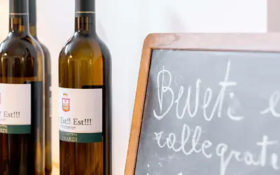 Bottiglie di vino "Est!! Est!!!" in primo piano, con una lavagna che invita a "Bevete e rallegratevi".