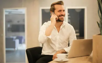 Uśmiechnięty mężczyzna w białej koszuli rozmawia przez telefon, siedząc przy biurku z laptopem. Może właśnie dowiaduje się, które banki dzwonią do pracodawcy.