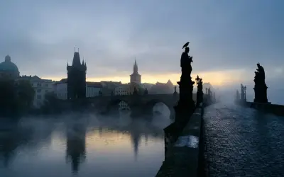 Mglisty poranek na praskim moście Karola. W oddali widać wieże i budynki, a na pierwszym planie kamienna balustrada i rzeźby.