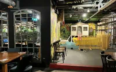 Warszawska restauracja z salą zabaw dla dzieci. Widać stoliki, krzesła i kolorowy plac zabaw z domkiem i zjeżdżalnią.