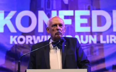 Janusz Korwin-Mikke, znany z kontrowersyjnych poglądów, przemawia na tle napisu "Konfederacja".