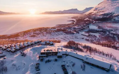 Vinterlandskap vid Kiruna med snötäckta fjäll, en frusen sjö och små stugor. Solen går ner och färgar himlen rosa.