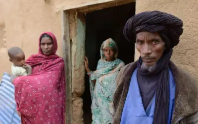 Ein Tuareg-Mann mit schwarzem Turban und ein Kind mit seiner Mutter vor einem Lehmhaus in der Sahara.