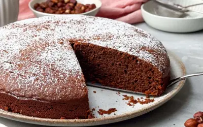Ein saftiger Nutella Kuchen, bestäubt mit Puderzucker, wartet darauf, angeschnitten zu werden. Daneben eine Schüssel mit Haselnüssen.