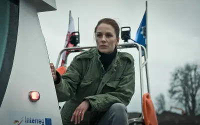 Katarzyna Wajda, aktorka, w zielonej kurtce, z polską flagą w tle, na tle pochmurnego nieba.