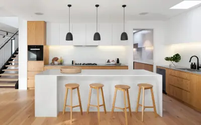 Moderna cocina blanca y madera clara. Isla central con taburetes, electrodomésticos integrados y suelo de madera.