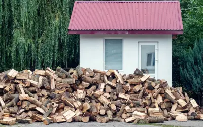 Duża sterta drewna opałowego przed białym budynkiem, z zielonym lasem w tle. Odległość budynku od lasu jest niewielka.