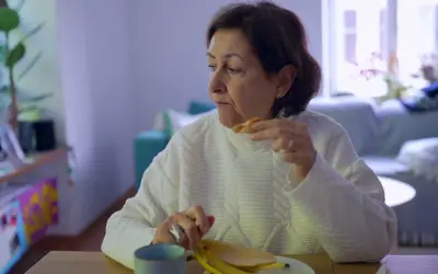 Kobieta w białym swetrze je banana, czując uczucie guli w gardle. Na stole kubek i talerz.