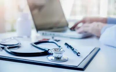 Stetoskop, długopis i notatnik na biurku lekarskim, w tle widać pracującego lekarza przy laptopie.