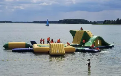 Rodzinna zabawa na wodnym placu zabaw na Mazurach. Dzieci w kamizelkach ratunkowych wspinają się i ślizgają po dmuchanych konstrukcjach.