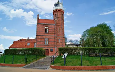Ceglana latarnia morska w Ustce z czerwoną dachówką i zielonymi żywopłotami.