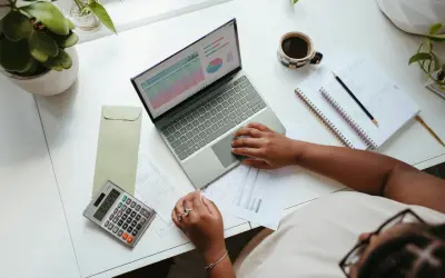 Biurko z laptopem, kalkulatorem, dokumentami i kawą, osoba pracuje nad finansami.