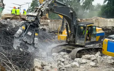 Maszyna budowlana wykonuje rozbiórkę budynku, krusząc beton i stal. W tle widać pracowników w kamizelkach odblaskowych.