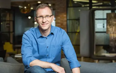 Marcin Iwuć, twórca "Finansowej fortecy", w niebieskiej koszuli siedzi na kanapie, uśmiechając się do obiektywu.