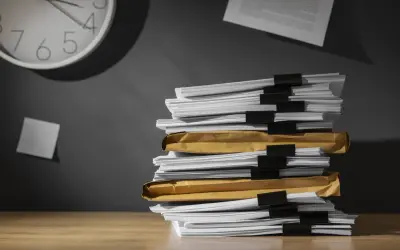 Una pila di documenti su una scrivania, con una nota appiccicata al muro e un orologio che segna il tempo. Chissà ogni quanto si aggiorna il dvr con tutto questo lavoro arretrato.