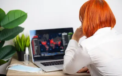 Kobieta z rudymi włosami patrzy na ekran laptopa z mapą świata i danymi. Obok leży maseczka i płyn do dezynfekcji.