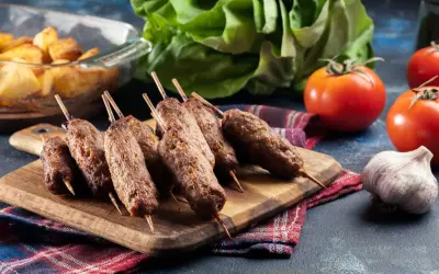 Grillowane ćevapi na drewnianej desce, obok pomidorów, czosnku i pieczonych ziemniaków.