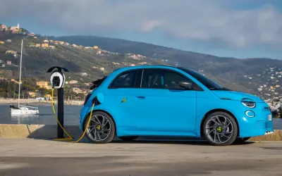 Abarth EV azul cargando junto al mar, con un velero y colinas al fondo.