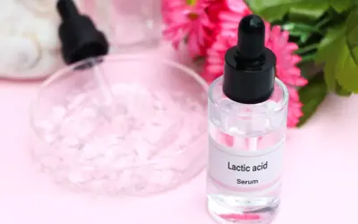 Serum z kwasem mlekowym w szklanej buteleczce z pipetą, obok różowych kwiatów i naczynia laboratoryjnego.