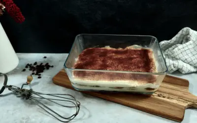 Un délicieux tiramisu 6 personnes, prêt à être dégusté, saupoudré de cacao. Un fouet ancien et des grains de café complètent la scène.
