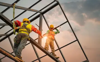 Dwóch budowlańców w kaskach i kamizelkach pracujących na wysokości. Za stan bezpieczeństwa i higieny pracy w zakładzie odpowiada każdy pracownik.