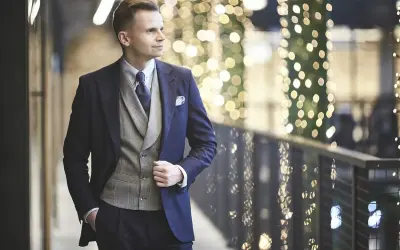 Elegancki mężczyzna w granatowym garniturze i kamizelce, gotowy na wieczór w stylu cocktail attire, na tle migoczących świateł.
