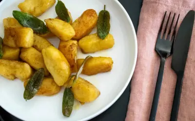 Goldbraune salbei gnocchi auf weißem Teller, garniert mit frischen Salbeiblättern. Daneben liegen schwarzes Besteck auf rosafarbenem Stoff.