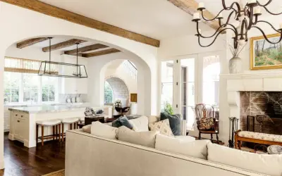 A cozy living room with a fireplace and kitchen, featuring exposed wooden beams and arched doorways, embodying a warm mediterranean interior design.