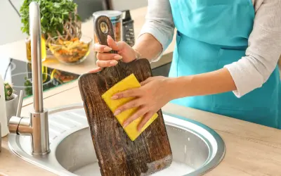Frau reinigt ein Holz-Schneidebrett mit einem gelben Schwamm in der Spüle. Backpulver hilft, hartnäckige Flecken zu entfernen.
