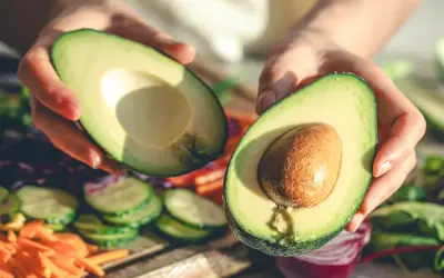Manos sostienen un aguacate cortado por la mitad, listo para disfrutar. Es la temporada del aguacate en España, con verduras frescas de fondo.