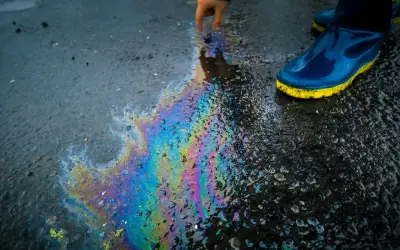 Tęczowa plama oleju na mokrym asfalcie, obok buta w kaloszu.