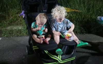 Two young children sit in a double stroller, eating corn from cups.