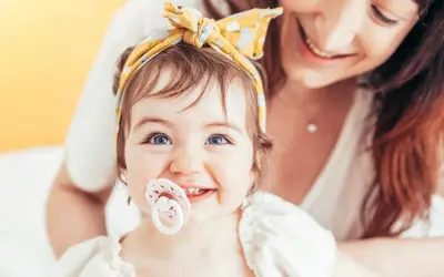 A baby with bright blue eyes and a yellow headband smiles, her passy teeth peeking out from behind a pacifier. Her mother smiles in the background.