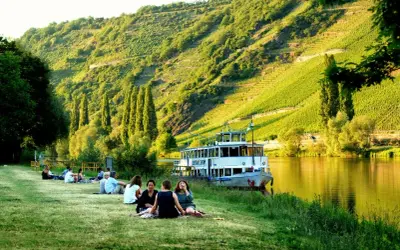 Entspannte Menschen genießen die Sonne am Ufer der Mosel. Ein Ausflugsschiff liegt vor, im Hintergrund steile Weinberge. Perfekte Mosel Ausflugsziele.
