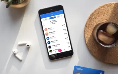 Telefon z aplikacją Revolut, pokazujący wydatki i opcje, np. revolut ubezpieczenie podróżne. Obok słuchawki i drink.