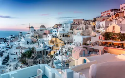 Santorini nocą, z białymi domkami, wiatrakami i grecką flagą, w blasku zachodzącego słońca.