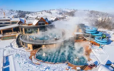 Gorący aquapark w górach, otoczony śniegiem, z parującymi basenami i niebieskimi zjeżdżalniami.