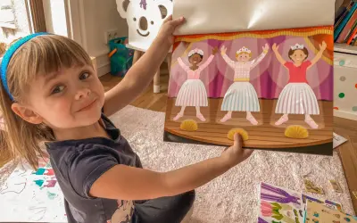 Uśmiechnięta dziewczynka pokazuje książeczkę z naklejkami baletnic. Idealny prezent na zajączka dla 3 latka, który uwielbia tańczyć.