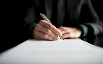 Dłoń pisze piórem na dokumencie. Może to być dziennik budowy, przerwa we wpisach lub ważny kontrakt.