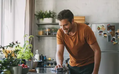 Uomo sorridente pulisce il piano cottura con un panno, mentre sul bancone ci sono piante aromatiche e pomodorini. Forse usa cristalli di soda per lucidare.