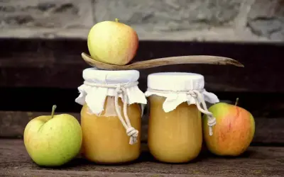Deux pots de compote de pommes faits maison, décorés de tissus et de cordes, avec des pommes fraîches et une cuillère en bois.