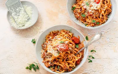 Dwa talerze spaghetti bolognese, posypane parmezanem. Obok miseczka z tartym serem i tarka. Długie gotowanie sosu to klucz do głębi smaku.