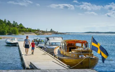 En kvinna och ett barn går på en brygga med båtar och en svensk flagga. En perfekt bild av en semester i Sverige.