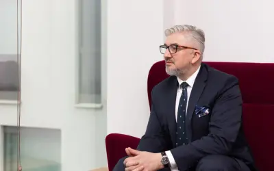 Mężczyzna w garniturze i okularach siedzi na czerwonym fotelu, patrząc w bok.